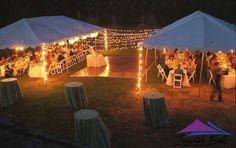 a group of people sitting at tables under white tents with string lights on the ground