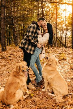 a man and woman standing next to two dogs in the woods with leaves on the ground