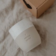 a white coffee mug sitting on top of a bed next to a box with the word coffee lover written on it