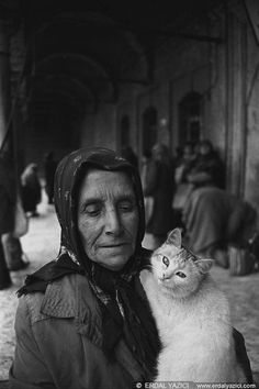 an old woman holding a white cat in her arms and looking at the camera, with other people standing around