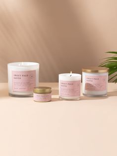 three candles with pink labels on them sitting next to a potted plant and palm leaf