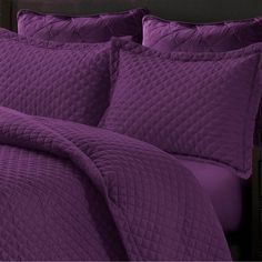 a bed with purple comforters and pillows on top of it, in front of a black headboard