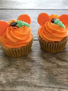 two cupcakes with orange frosting and mickey mouse ears on them sitting on a wooden table