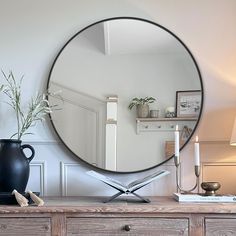 a round mirror sitting on top of a wooden dresser next to a vase and lamp