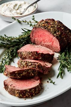 sliced meat on a white plate with herbs and dip in the bowl next to it