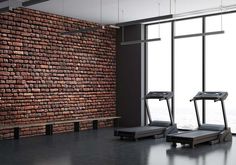 there are two treadmills next to a brick wall in an empty room with large windows