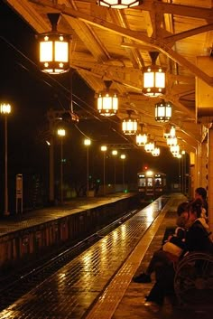 two people sitting on the side of a train platform at night with lights hanging above them