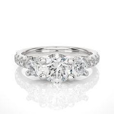 "This stunning brilliant trellis three stone engagement ring is a masterpiece of fine craftsmanship and exquisite design, perfectly combining the beauty of a 1.20+ carat Round lab-grown diamond with the sparkle of three stone diamonds. The Round diamond is of exceptional quality, with brilliant cut facets that maximize its natural radiance and fire. The diamond is set in a three stone shank design, which adds depth and dimension to the ring by framing the center stones appear to float invisibly above the surface of the ring. The ring's shank is also set with pave diamonds, which create a glittering band of brilliance that draws the eye toward the center stone. Each diamond is expertly set in a shared prong setting, which ensures that each stone is securely held in place while allowing maxi Luxury Three Stone Round Cut Diamond Ring, Gia Certified Round Cut Promise Ring, Elegant Three Stone Promise Ring, Three Stone Platinum Round Rings, Platinum Three Stone Round Rings, Luxury Three Stone 14k White Gold Rings, Luxury Three Stone Round Cut Wedding Ring, Timeless Three Stone Diamond Ring For Anniversary, Luxury Three-stone Round Cut Wedding Ring