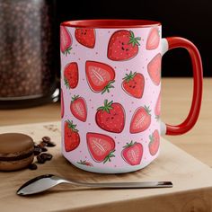 a red and white coffee mug with strawberries on it sitting next to a cookie