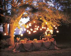 an outdoor dinner table under a tree with lanterns on the branches and lights hanging from it