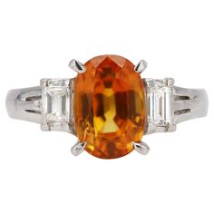 A vivacious and vivid orange sapphire gemstone engagement ring that offers the opportunity to own a rare, vintage heritage piece. For a non-traditional bridal ring, this fiery, deeply saturated orange gem is perfectly placed in a platinum setting shouldered by a single emerald cut diamond accent. Weighing in at a total of 3 carats, this vintage estate treasure is waiting for you.   Condition: Very good Era: Vintage Year: 1980s Metal: Platinum Gemstone: Sapphire Carat Weight: 2.93 Cut: Oval Color Luxury Orange Ring With Prong Setting, Luxury Orange Rings With Prong Setting, Orange Diamond Ring With Prong Setting, Luxury Vintage Orange Rings, Luxury Orange Yellow Sapphire Ring, Custom Ring Box, Orange Gem, Orange Sapphire, Gemstone Engagement