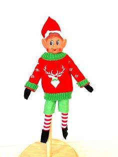 an elf doll is standing on top of a piece of wood and wearing a red sweater