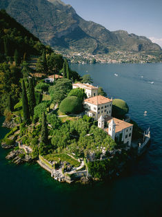 an island in the middle of water surrounded by trees and buildings with mountains in the background