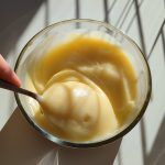 a person holding a spoon in a glass bowl filled with orange liquid and sauce on a white surface