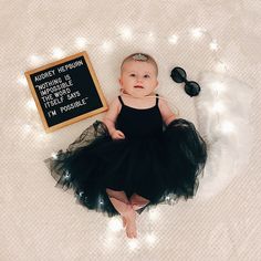 a baby wearing a black dress sitting on top of a white blanket next to a chalkboard