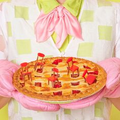 a woman in pink gloves holding a pie with red decorations on it's crust