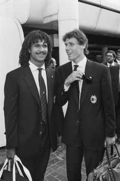 two men in suits standing next to each other holding bags and smiling at the camera