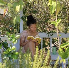 a woman sitting on a bench reading a book in the shade by some bushes and trees