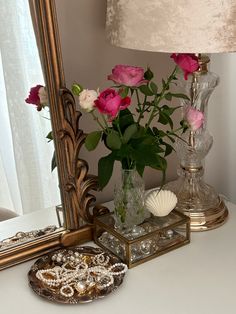 pink roses in a vase on a table next to a mirror and seashells