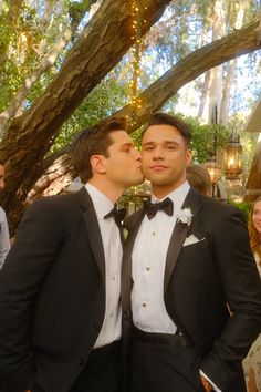 two men in tuxedos standing next to each other under a tree with lights