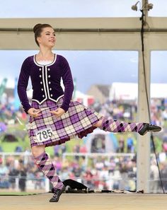 a woman in a kilt is dancing on the stage with people watching behind her