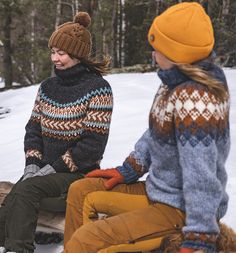 two women sitting on a log talking to each other in the snow with trees behind them