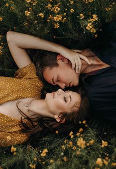 a man and woman laying on the ground in front of yellow flowers with their heads touching each other