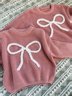 two knitted sweaters with bows on them are laying on a patterned surface, one is pink and the other is white