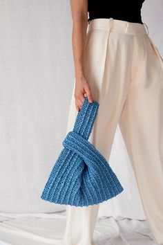 a woman holding a blue knitted purse in her right hand and wearing white pants