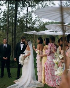 a bride and groom standing under an umbrella in the grass with other people around them