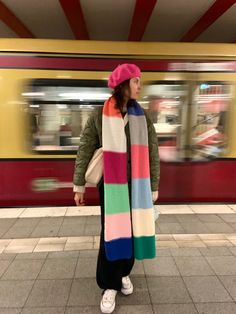 a woman wearing a multicolored scarf and hat stands in front of a subway train