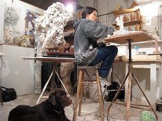 a woman sitting at a table with a dog in front of her and she is working on a sculpture