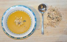 a bowl of carrot soup next to a spoon and granola on a wooden table