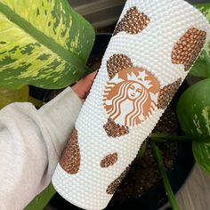 someone holding up a starbucks coffee cup decorated with gold and white sequins in front of a potted plant