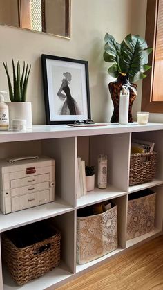 a white shelf with baskets and pictures on it next to a plant in a vase