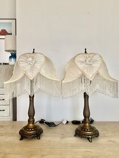 two lamps with fringed shades on them sitting on a wooden table in front of a white wall