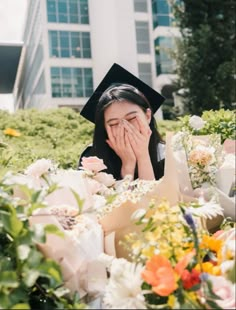 a graduate covers her face as she sits among flowers