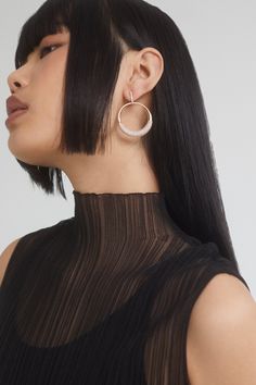 a woman with long black hair wearing large hoop earrings and a sheer top, looking off to the side