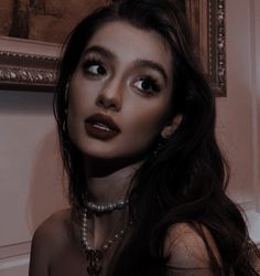 a woman with long dark hair and pearls on her neck posing in front of a painting