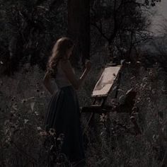 a woman standing in the woods with an easel next to her and looking at it