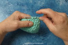 two hands are crocheting the end of a small green object on a blue surface