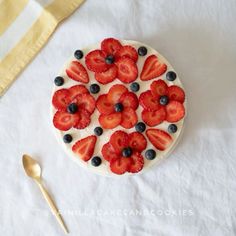 a white cake with strawberries and blueberries on it next to a gold spoon