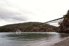 a large bridge spanning over a river next to a lush green forest covered hillside on a cloudy day