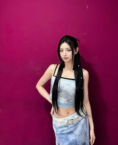 a woman with long black hair standing in front of a pink wall wearing a skirt