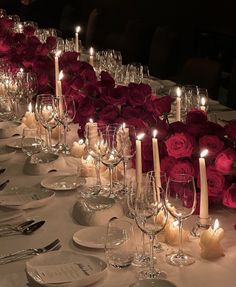 a long table is set with wine glasses, plates and candles in front of red roses