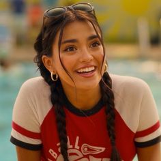 a woman with braids and sunglasses smiling at the camera while sitting by a swimming pool