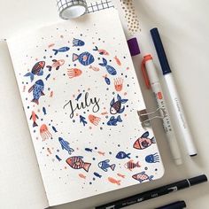 an open notebook with the word july written on it next to some pens and markers