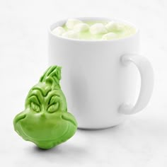 a mug with marshmallows in it next to a green creature head