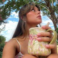 a woman sitting under a tree drinking out of a coffee cup with her eyes closed