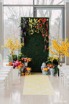 an indoor ceremony with flowers and greenery on the wall, along with white chairs